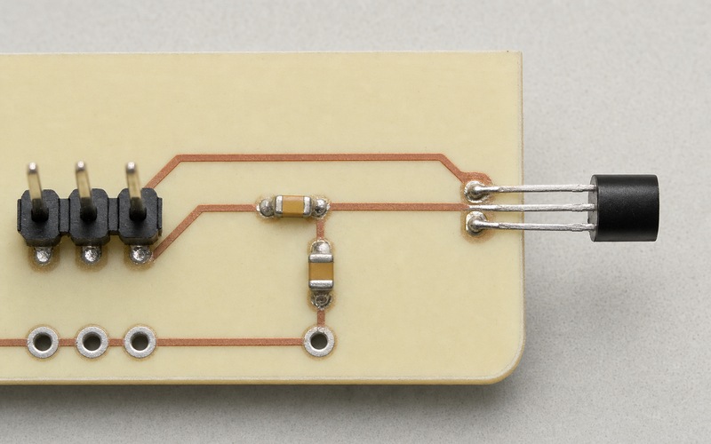 Draufsicht auf eine cremefarbene Platine mit TO-92-Temperatursensor am Außenrand und kurzer Kupferleitung durch ein kleines RC-Filter